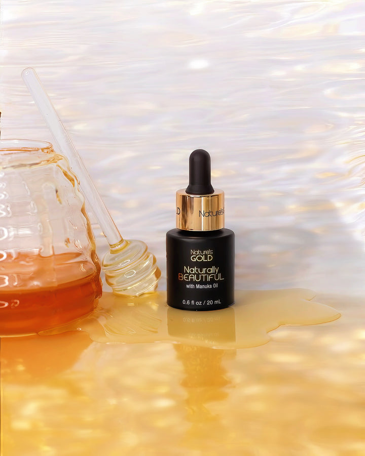 Bottle of Nature's Gold Naturally Beautiful with Manuka Oil on pool of honey and a glass honey spoon and honey jar at the sides on bright yellow and white background