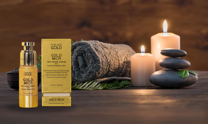 Bottle and Box of Nature's Gold Gold Mica Serum on a wooden table with a towel, candles and pebbles in a dark room